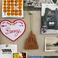 S.R. Air freshener on a pin up wall with various ephemera pinned next to it including an embroidered heart that says "My One True love PENNY". The S.R. Air freshener is mountain shaped.