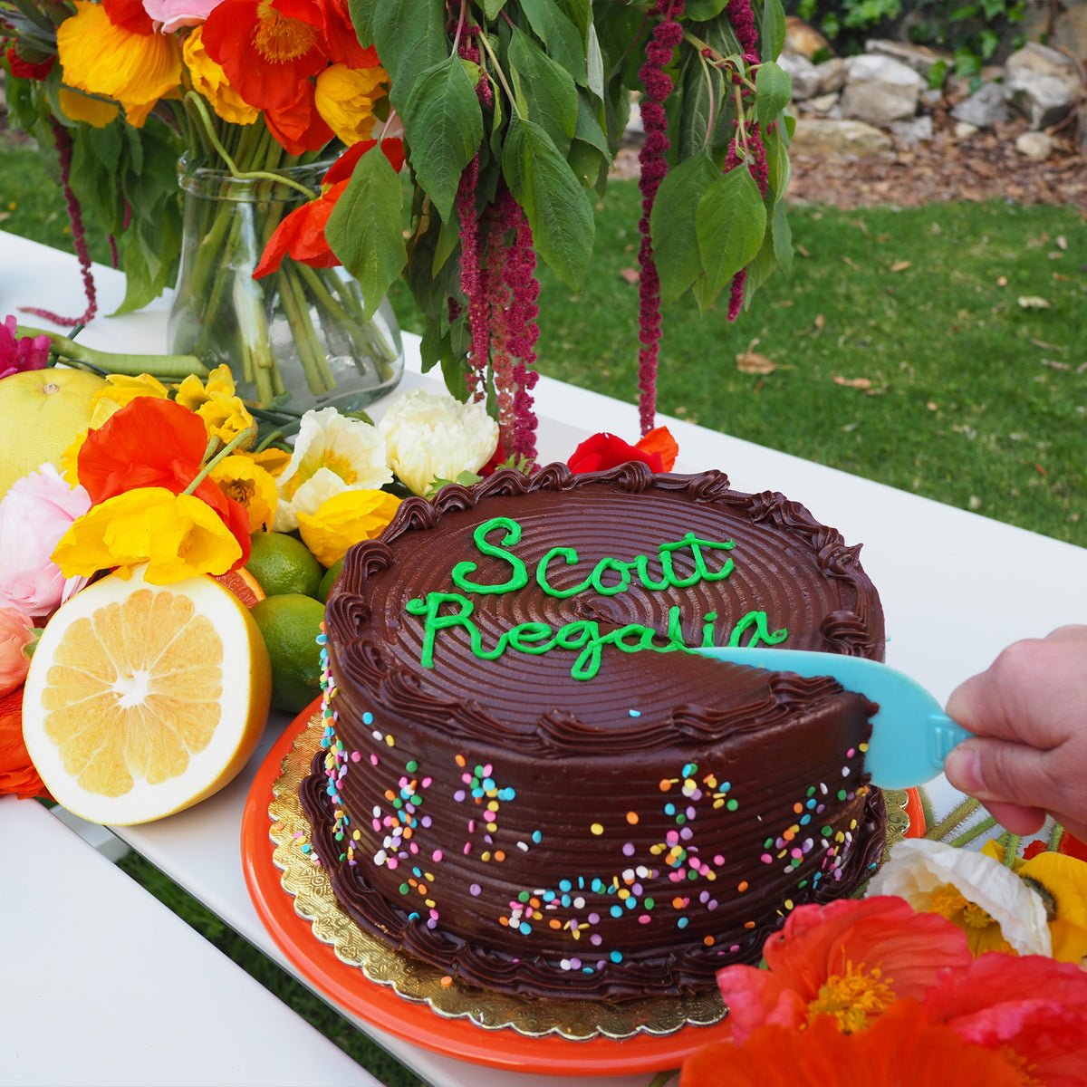 A chocolate cake with green frosted writing that says "Scout Regalia" being sliced next to flowers on a white table.
