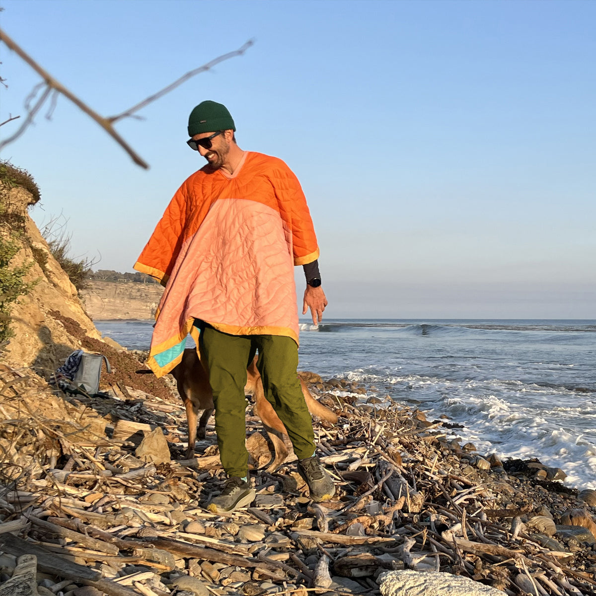 S.R. Anywear Blanket in Sunset Orange. Man wearing an orange and pink poncho on the beach standing on driftwood