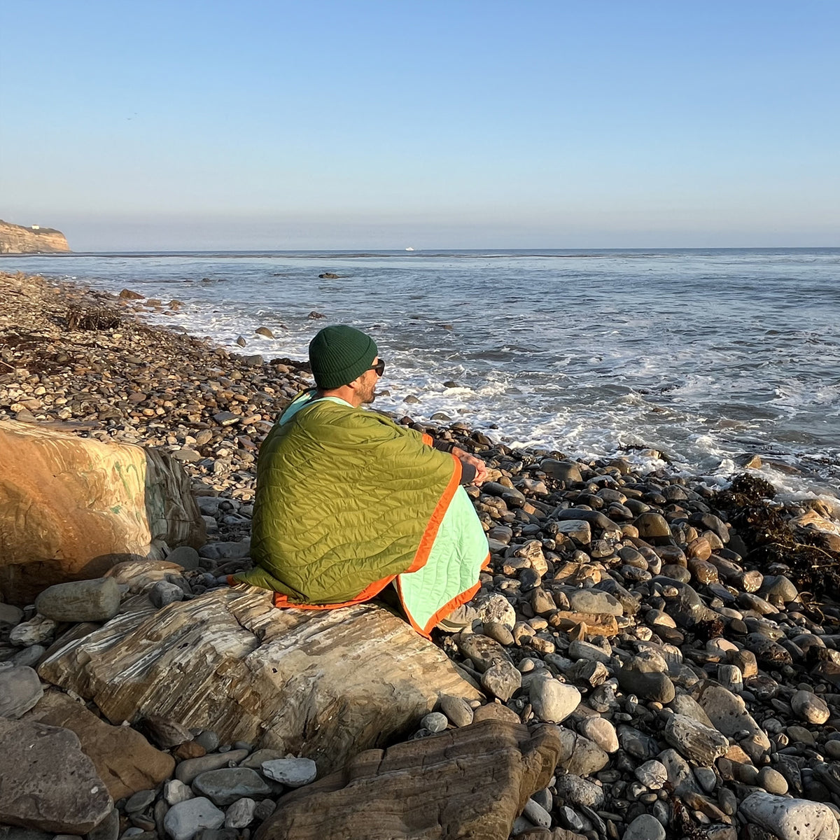 S.R. Anywear Blanket in Ocean Kelp. A man wrapped in a green poncho sitting on a rock overlooking the ocean