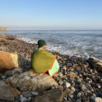 S.R. Anywear Blanket in Ocean Kelp. A man wrapped in a green poncho sitting on a rock overlooking the ocean