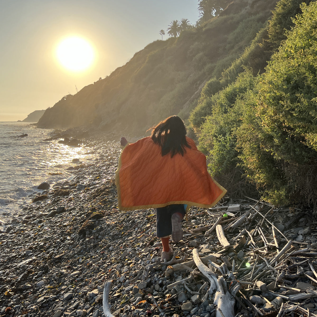 S.R. Anywear Blanket in Sunset Orange. A woman running away with an orange blanket on the beach