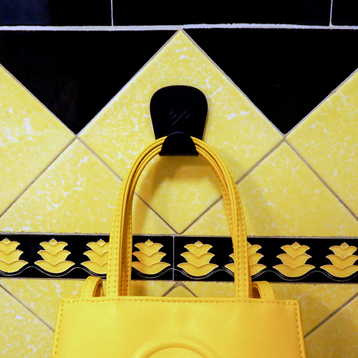 S.R. Anywhere Hook in Matte Black on a black and yellow tile wall, holding a yellow handbag