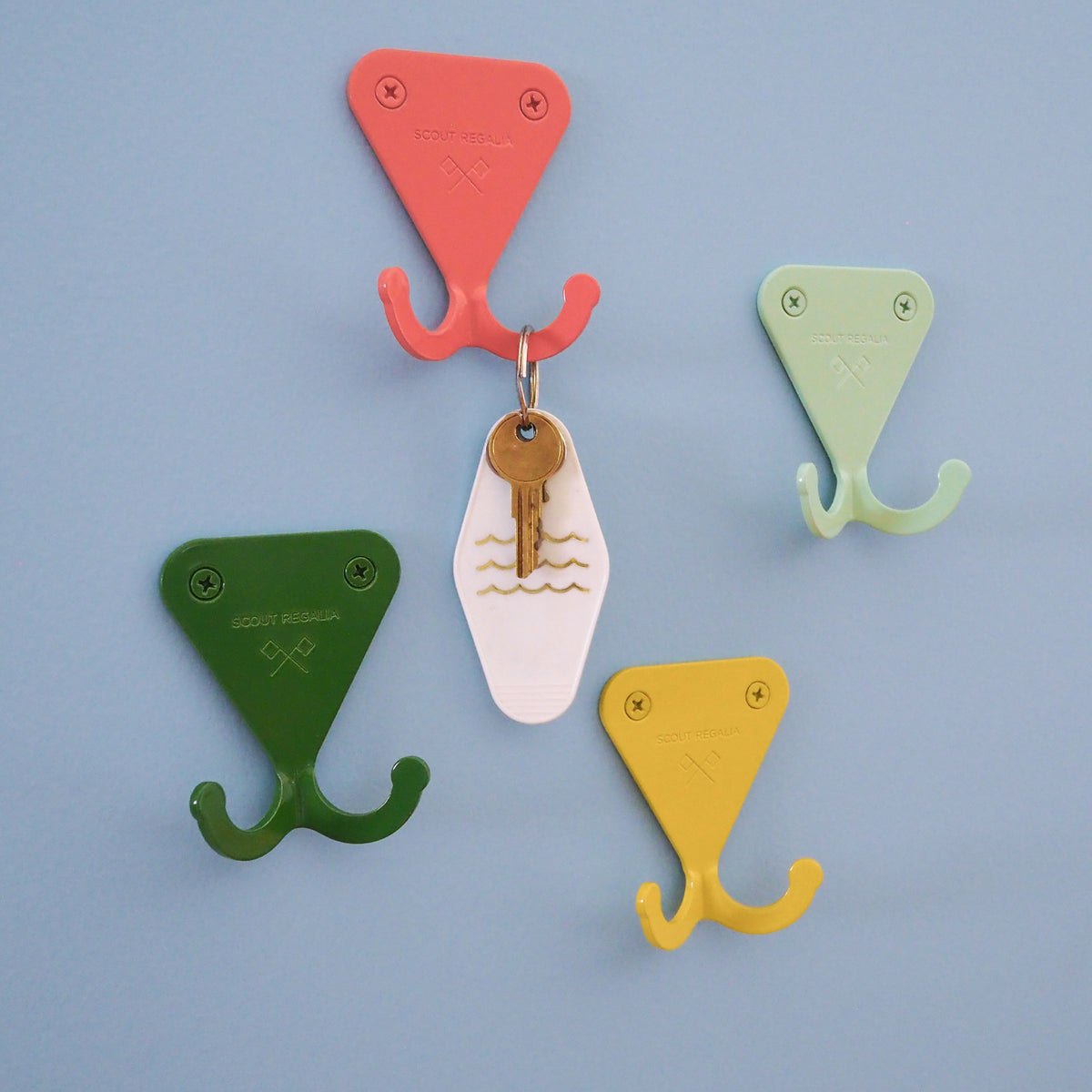 Salmon Pink, Mint Green, Fern Green, and Citrus Yellow S.R. Wall Hooks on a blue wall. The Salmon Pink S.R. Wall Hook is holding a key on a white keychain.