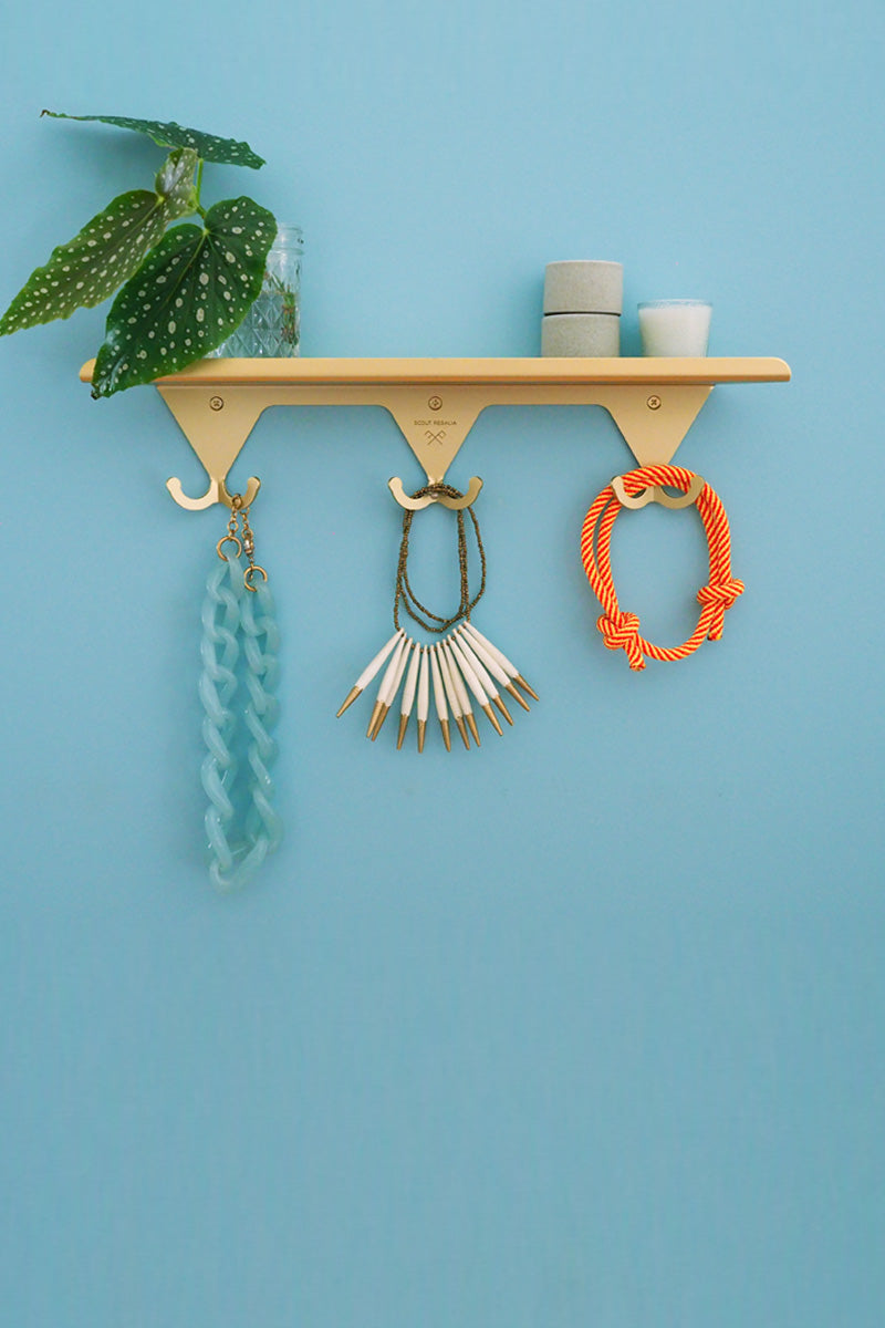 A Brass Scout Regalia Wall Rack on blue background with various necklaces and bracelets hanging on the hooks, and greenery in a small vase with ceramic stack and candle on the shelf.