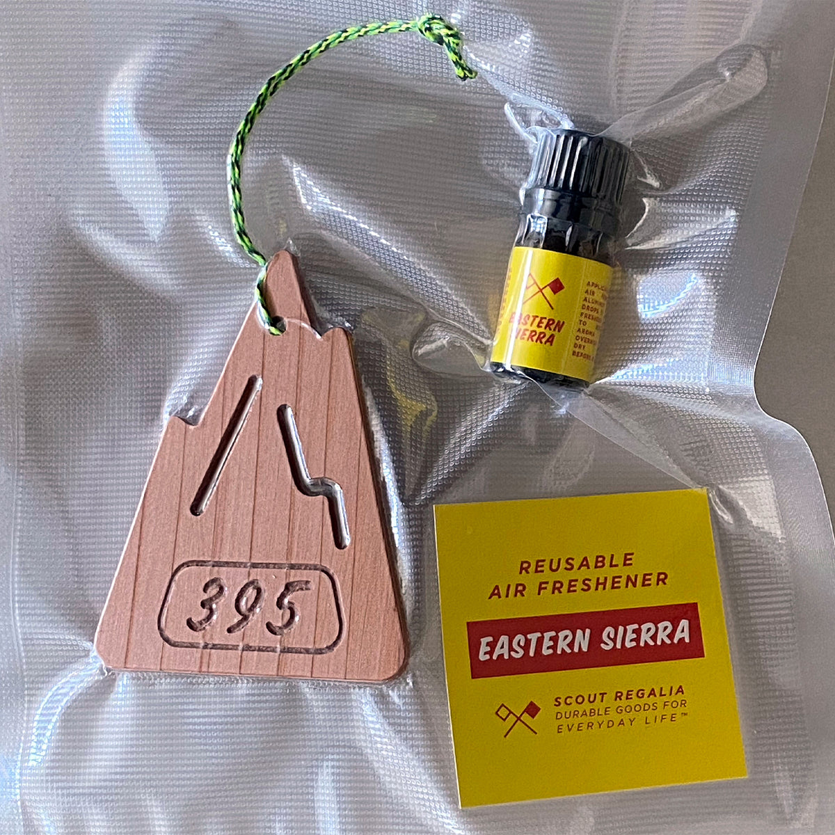 Packaging for the S.R. Air freshener. A wooden mountain shaped air freshener, yellow card that says" Reusable Air Freshener Eastern Sierra", and a vial of scented oil in an air sealed bag.