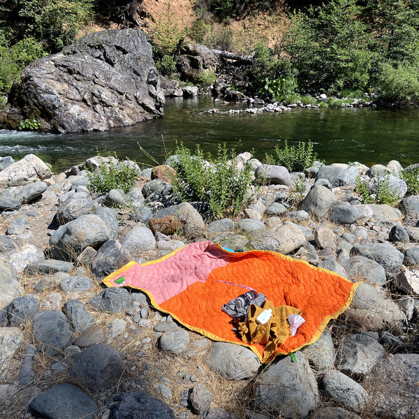 Orange and pink blanket with water bottle and towel on rocks next to a river