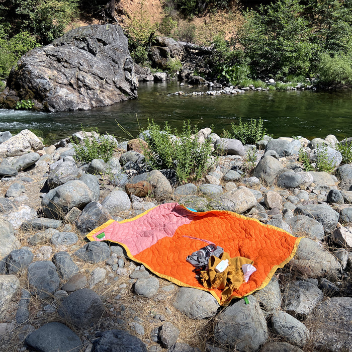 S.R. Anywear Blanket in Sunset Orange.  An orange and pink blanket with water bottle and towel on rocks next to a river