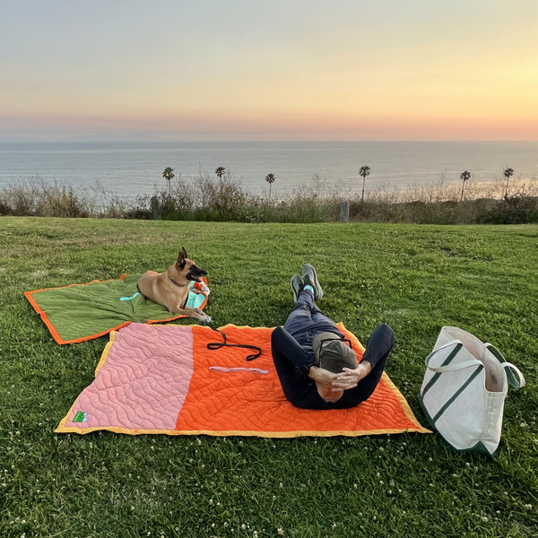 S.R. Anywear Blanket in Sunset Orange and Ocean Kelp. Man and dog laying on two blankets on the grass overlooking ocean view.