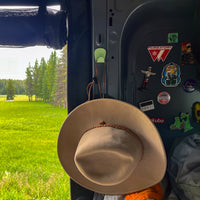 Mint green S.R. Anywhere Hook on the inside of a grey van with a brown hat hanging off it. Green field in the background with various magnets on the inside of van.