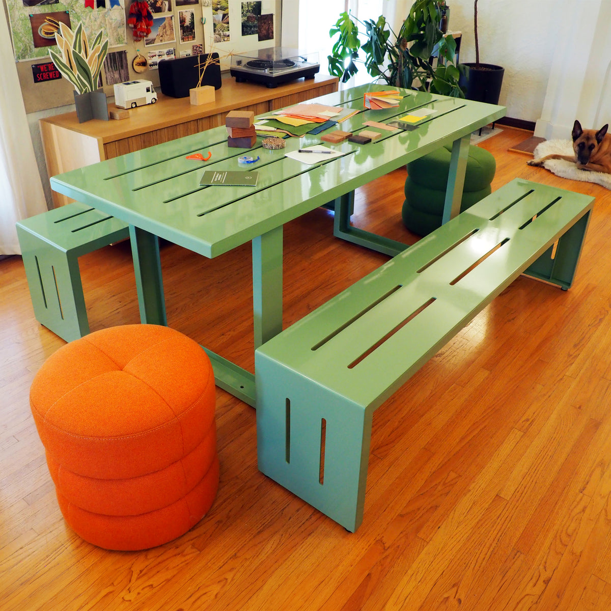 A pale green S.R. All Aluminum Table Set with material samples on the table. The S.R. All Aluminum Table Set is in a room with hardwood floors and an orange S.R. Pouf on the end of the table.