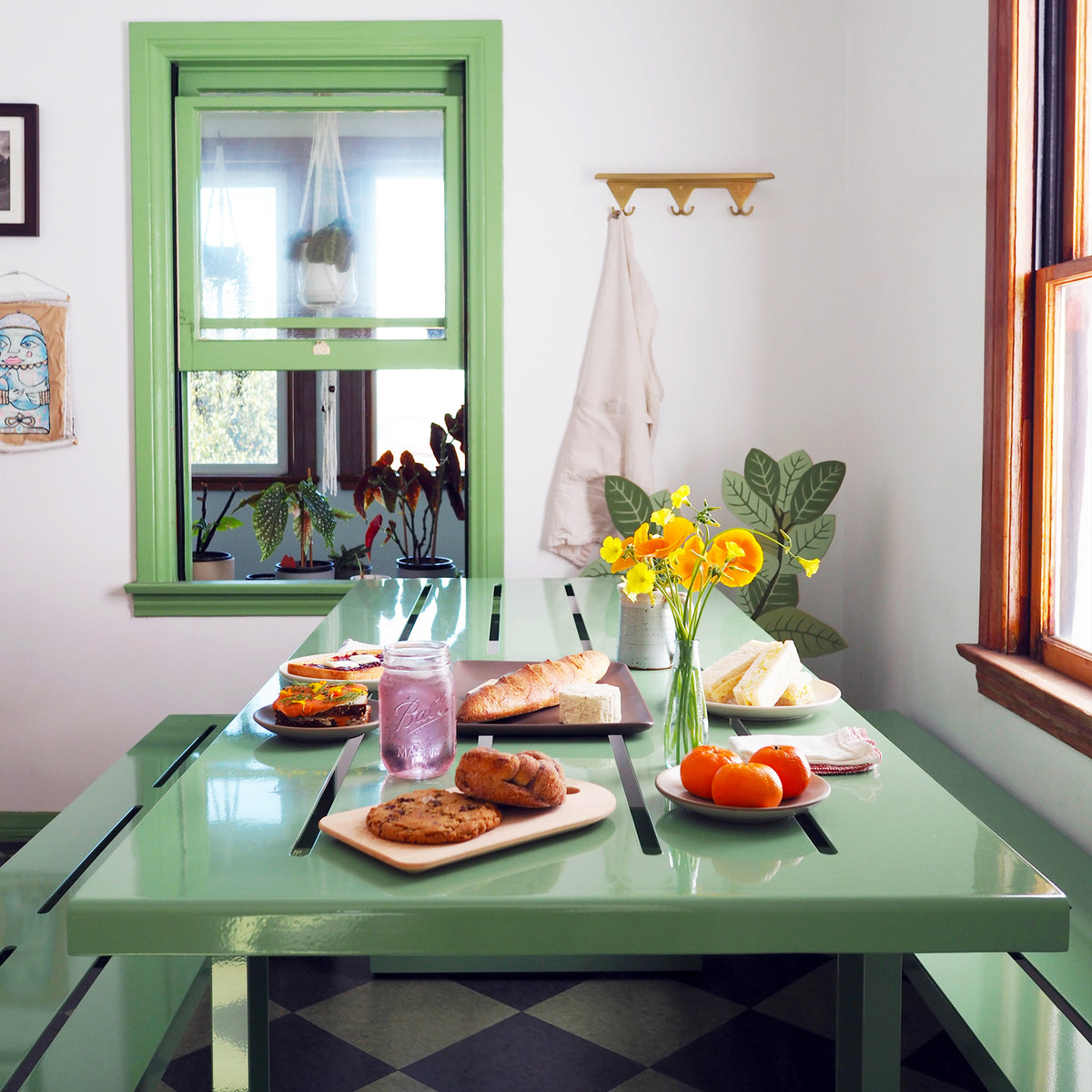 A pale green S.R. All Aluminum Table Set with pastries, fruit, and bread on the table top with a vase of orange flowers. The table is in a room with a green framed window and a brass S.R. Wall Rack with an apron on the wall.