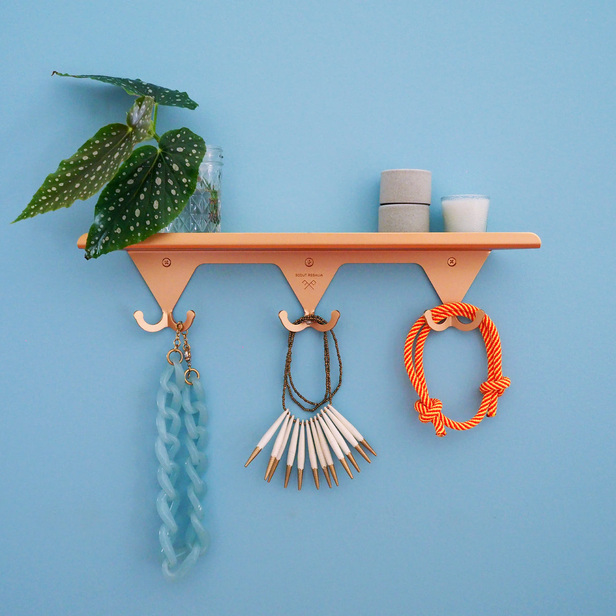 A Brass S.R. Wall Rack on blue background with various necklaces and bracelets hanging on the hooks, and greenery in a small vase with ceramic stack and candle on the shelf.