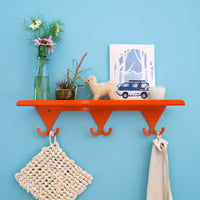 Bright Orange S.R. Wall Rack with flower vases, a small wool alpaca toy, a card with a painted Delica van, and candle on shelf. A macrame potholder in ivory and linen apron hang on the hooks.